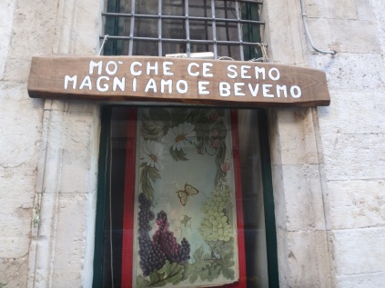 Translation: "GIA' CHE CI SIAMO, MANGIAMO E BEVIAMO" a Romanesco dialect. Which means "We're here, eating and drinking." (Sounds better in Italian.)